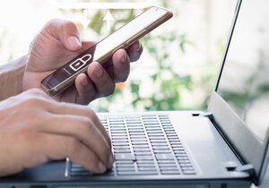 Zwei Hände, die recht ruht auf einer Laptoptastatur, die linke hält ein Smartphone